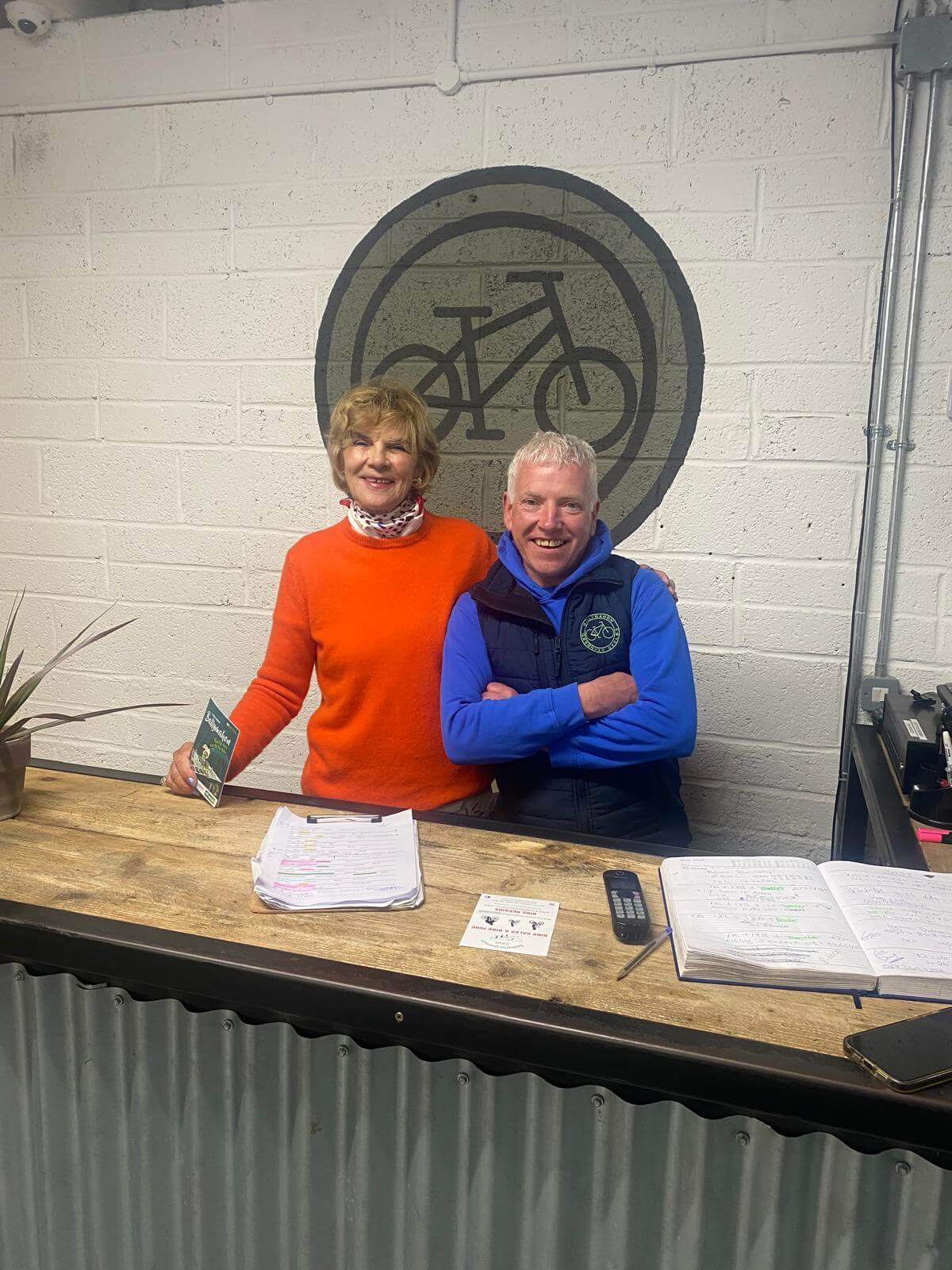 Two smiling people stand behind a counter in a bike shop. The woman wears an orange jumper and holds a leaflet; the man wears a blue waistcoat. Papers, a plant, and a notebook are on the counter. A bicycle logo is on the wall behind them.