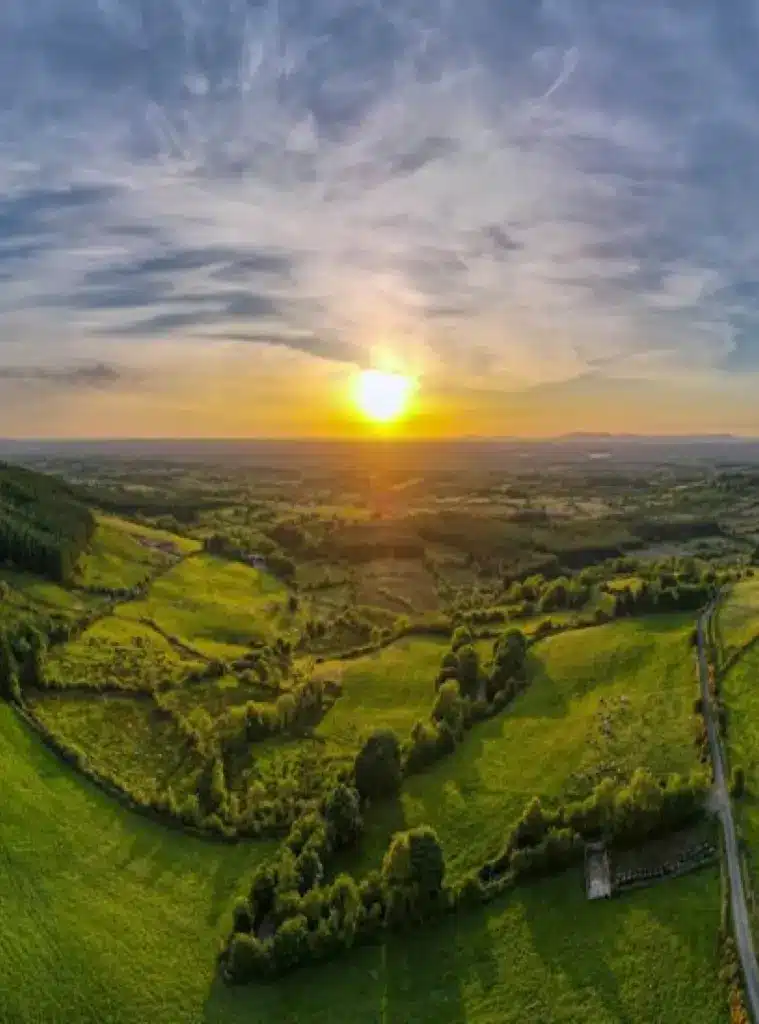 Aerial view of a lush green landscape with fields, trees, and hedgerows under a dramatic sky as the sun sets on the horizon, casting a warm golden light over the countryside.