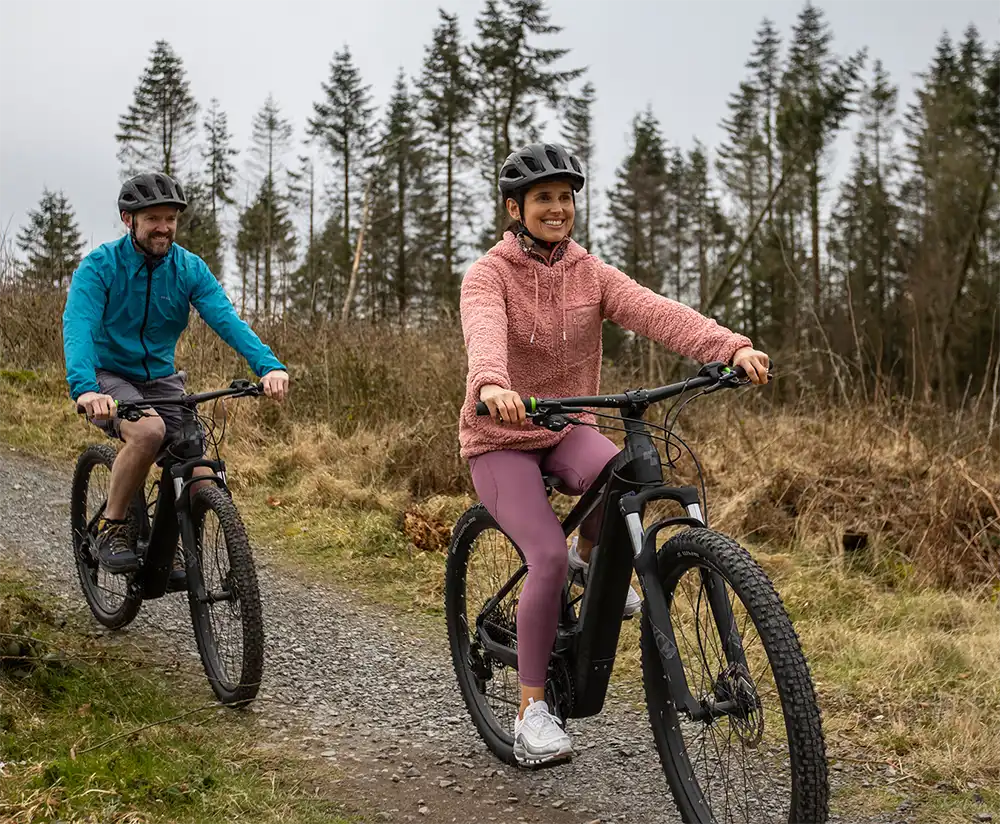 Two people wearing helmets ride electric bikes on a gravel path through a forested area. The person in front is smiling and wearing a pink jacket; the person behind is in a blue jacket. Trees and grass are in the background.