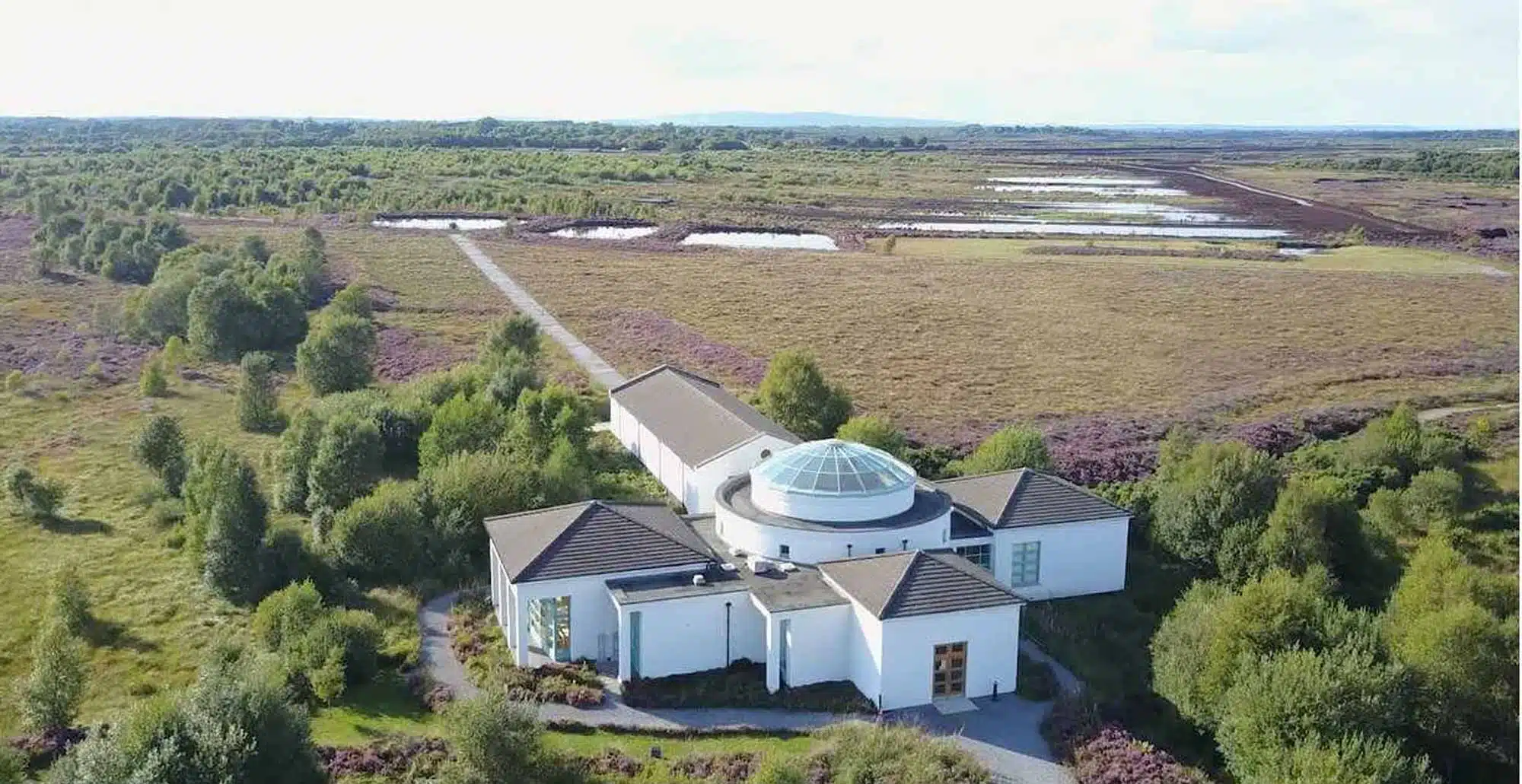 Aerial view of a modern white building with a glass dome roof, surrounded by trees and open fields with patches of water under a bright sky. A long path leads away from the building through the landscape.