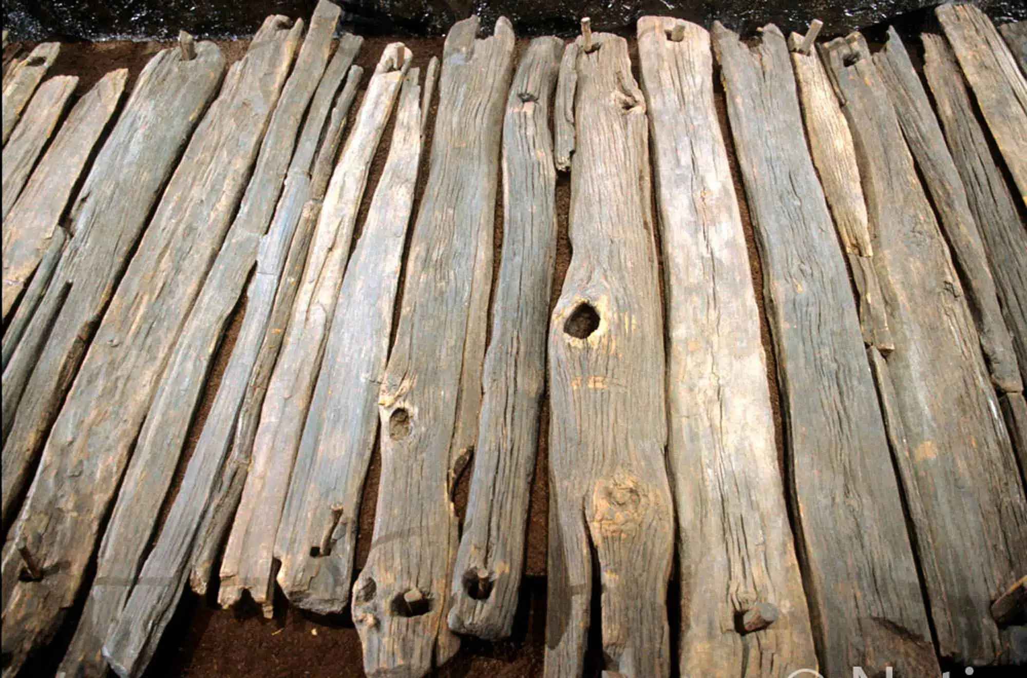 A close-up view of several weathered wooden planks laid side by side outdoors, forming a flat surface. The wood appears rough, aged, and uneven, with visible knots, cracks, and natural texture.