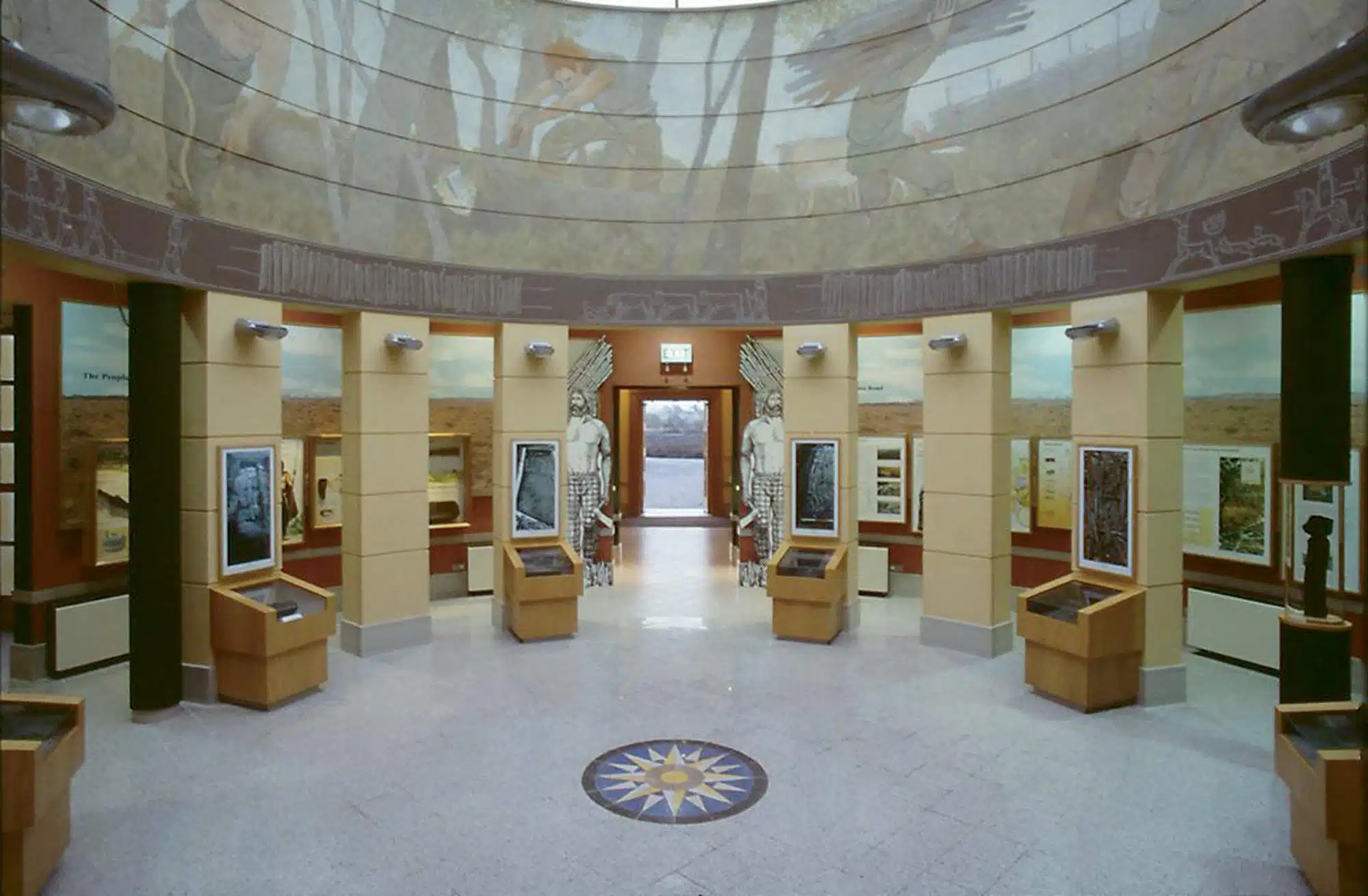 A circular museum exhibition room with display cases, information panels, and a compass rose on the floor. The domed ceiling is decorated with a mural of figures and landscape scenes. Natural light enters through glass doors at the far end.