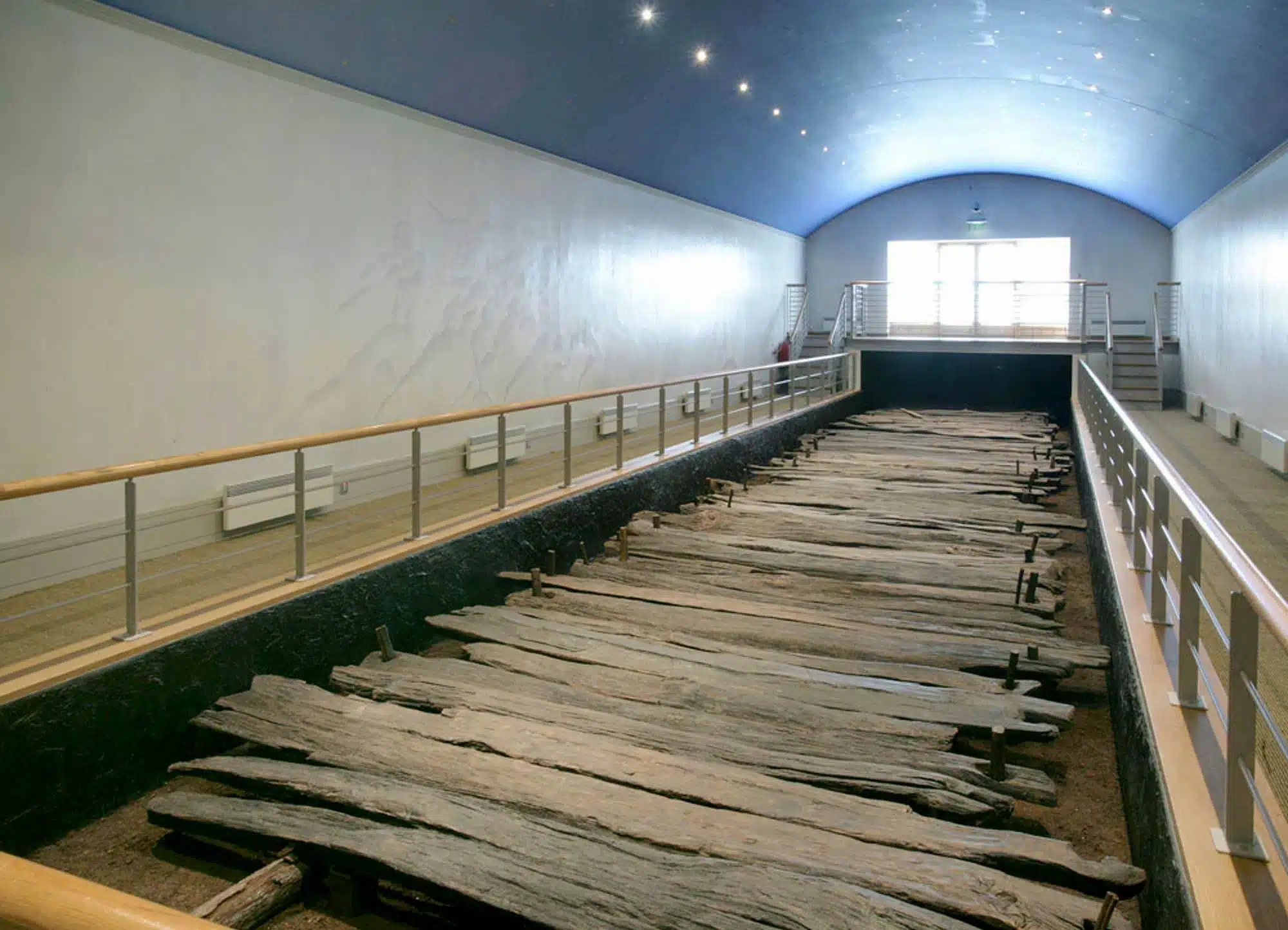 Large ancient wooden boat remains are displayed in a long, well-lit museum room with railings along both sides and a curved blue ceiling.