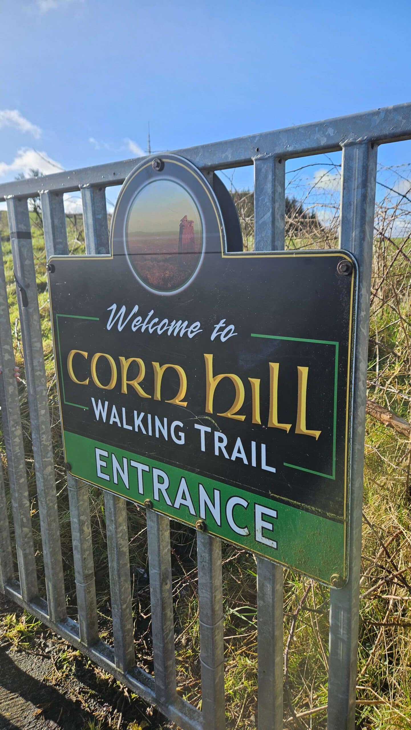 A metal gate with a sign reading “Welcome to Corn Hill Walking Trail Entrance” under a small image of a hill with a tower, set outdoors on a sunny day.