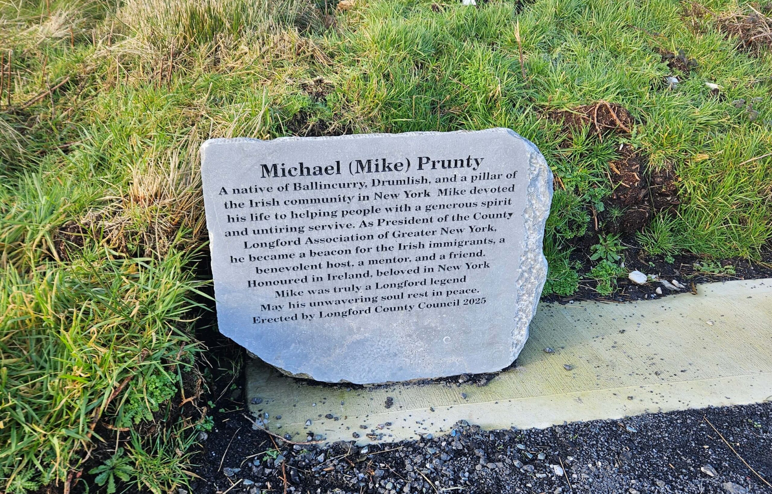A memorial stone set in grass honours Michael (Mike) Prunty with an inscription celebrating his dedication to the Irish community in New York and his generous spirit.