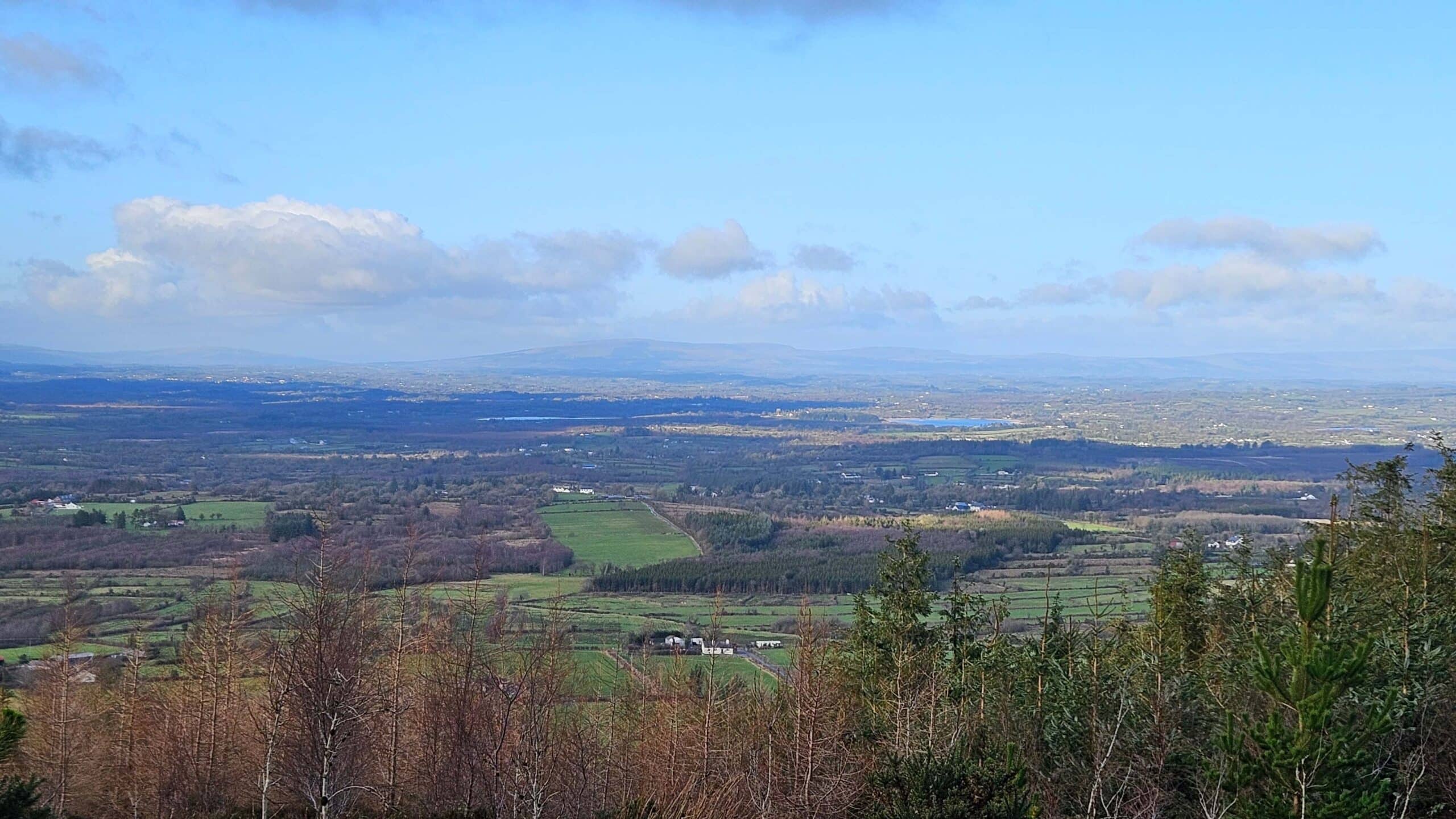 A panoramic view of a rural landscape with fields, scattered houses, and patches of woodland under a blue sky with scattered clouds; distant hills stretch across the horizon.