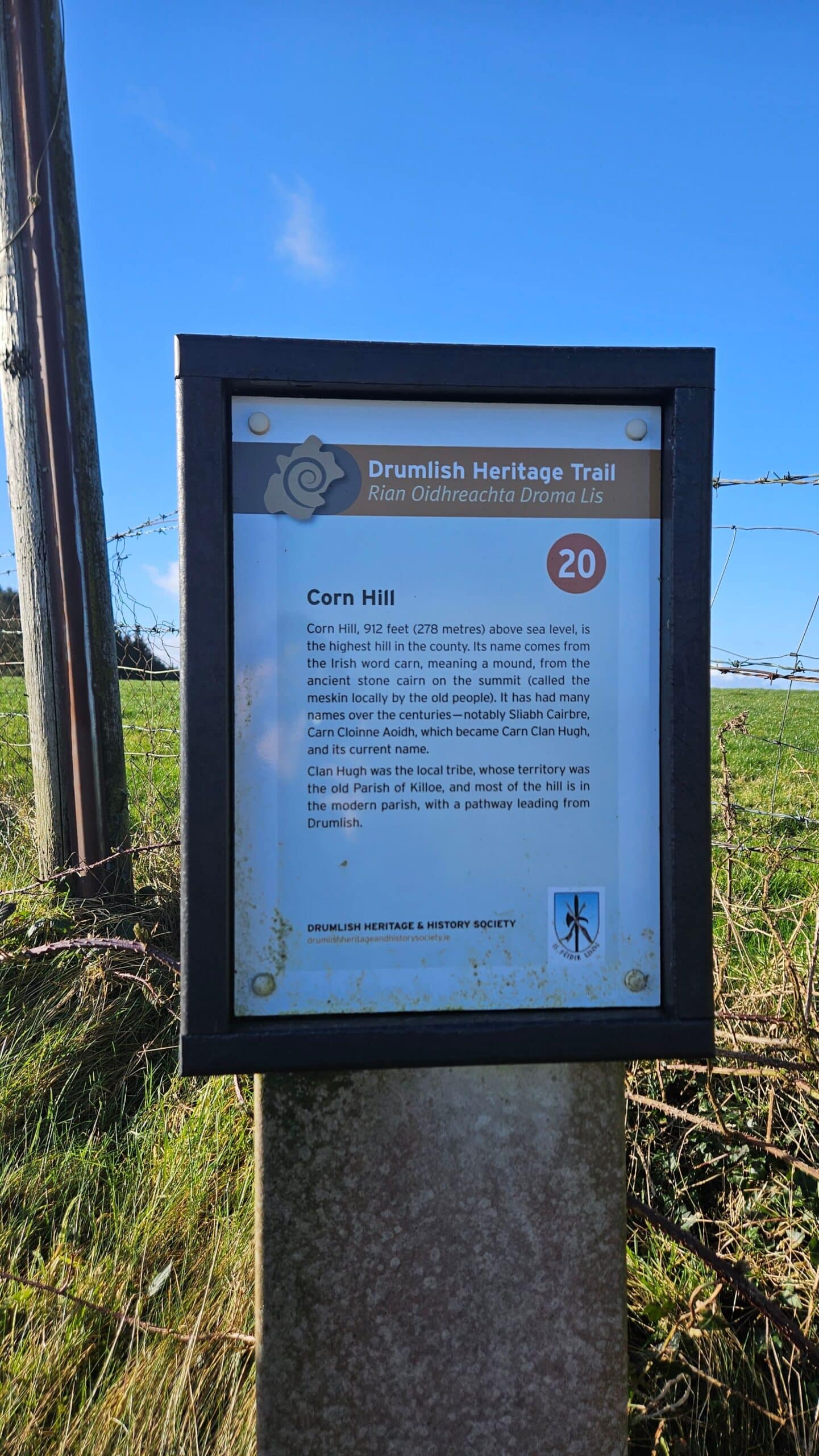 A signpost for the Drumlish Heritage Trail stands on grass by a barbed wire fence. It provides information about Corn Hill (altitude 942 feet) and its local history, with the number 20 in an orange circle at the top right-hand corner.