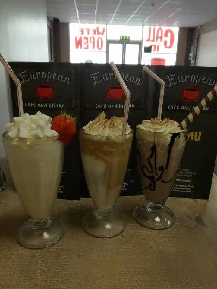 Three milkshakes—vanilla with marshmallows and a strawberry, caramel with whipped cream, and chocolate with whipped cream and a wafer stick—are on a counter in front of "European Café and Bistro" menus.