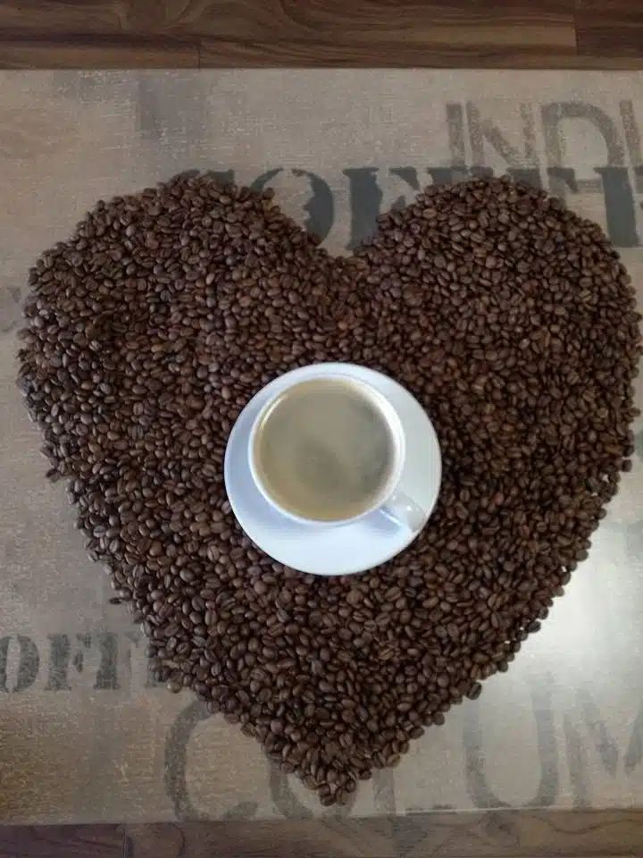 A white cup of coffee sits on a saucer placed at the centre of a heart shape made from coffee beans on a brown surface.