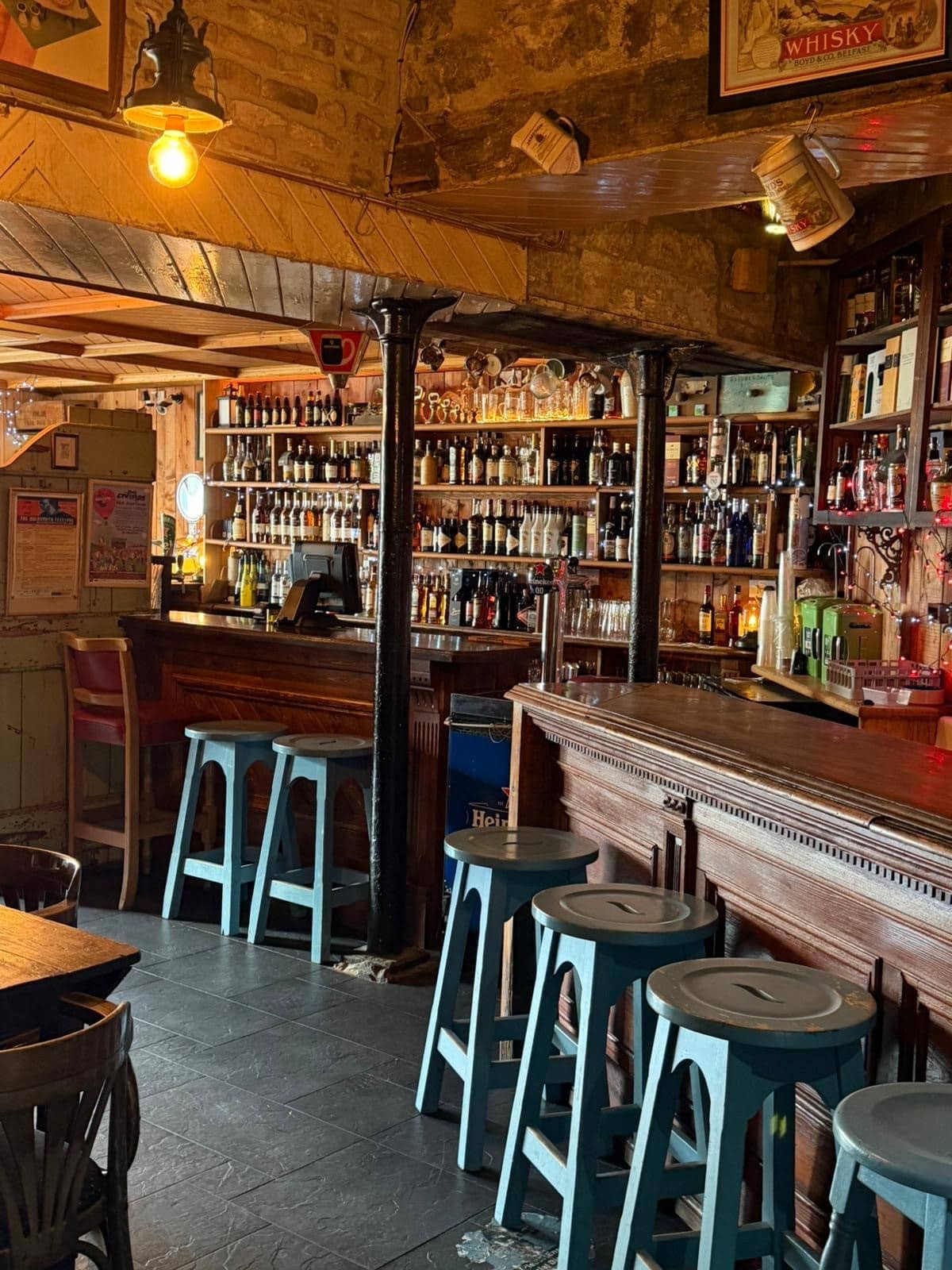 A cosy pub interior with wooden bar counters, shelves filled with bottles, blue bar stools, exposed stone walls, and warm lighting creating a welcoming atmosphere.