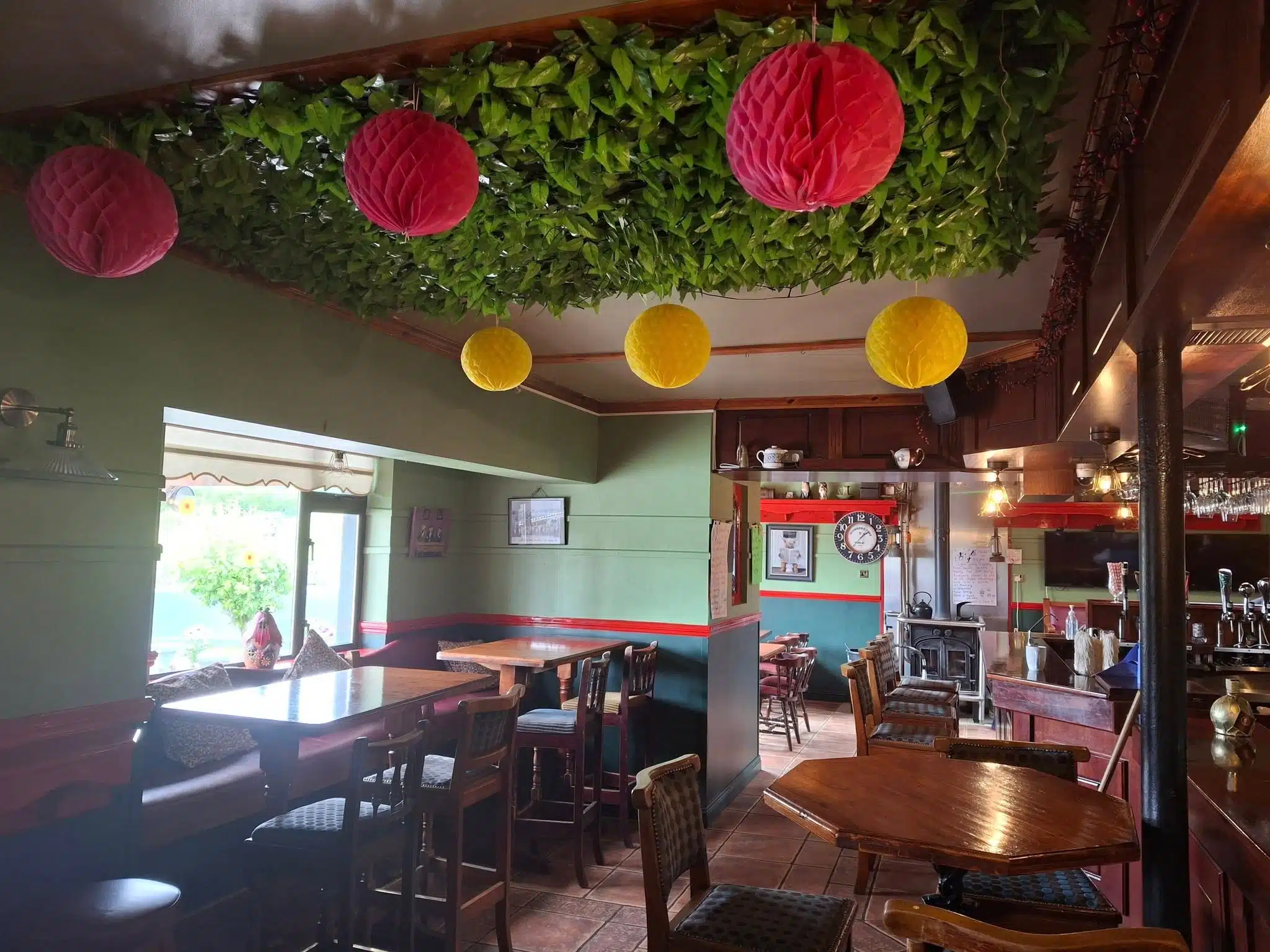 Cosy bar interior with wooden tables and chairs, leafy ceiling decor, and pink and yellow paper lanterns hanging. Large windows let in sunlight, creating a warm and inviting atmosphere.