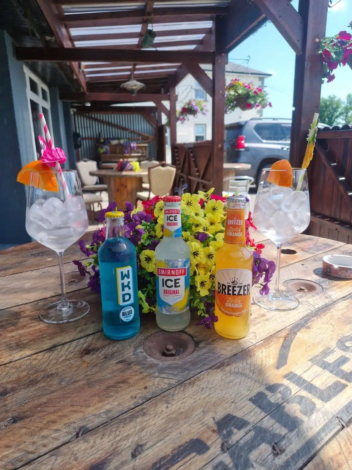 Three colourful bottled drinks (WKD Blue, Smirnoff Ice, and Breezer Orange) are placed on a rustic wooden table with two glasses of ice and slices of orange, surrounded by vibrant flowers in an outdoor seating area.