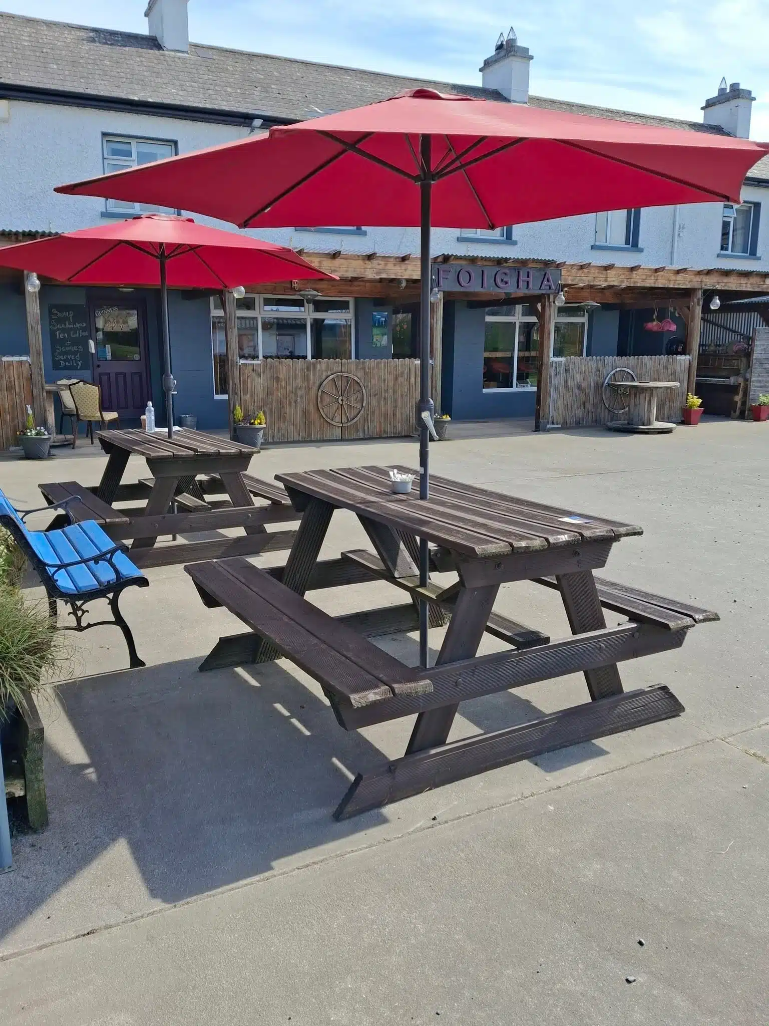 Wooden picnic tables with bright red umbrellas are set outside a rustic café with a wooden facade and a sign reading "FOICHE." A blue bench and flowerpots decorate the area on a sunny day.