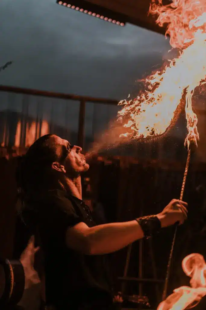 A performer in dark clothes and sunglasses blows a large plume of fire from their mouth whilst holding a lit torch, creating a striking scene against the dimly lit backdrop at a Sustainable Tourism Longford event.