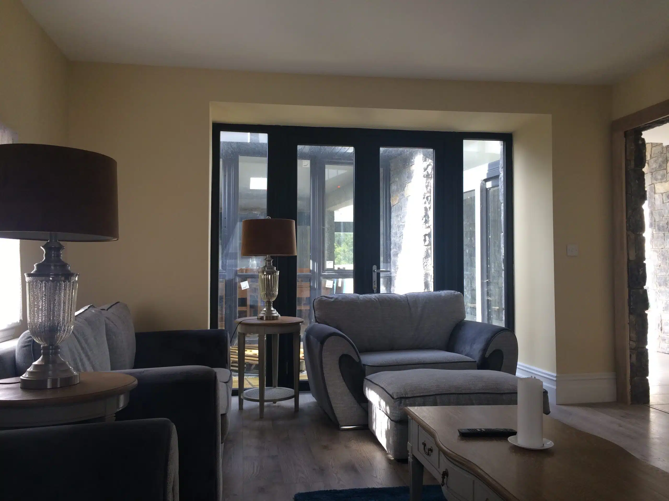 A cosy living room with grey sofas, two glass table lamps, a wooden coffee table with a candle and remote, and large glass doors letting in sunlight from an outdoor area.