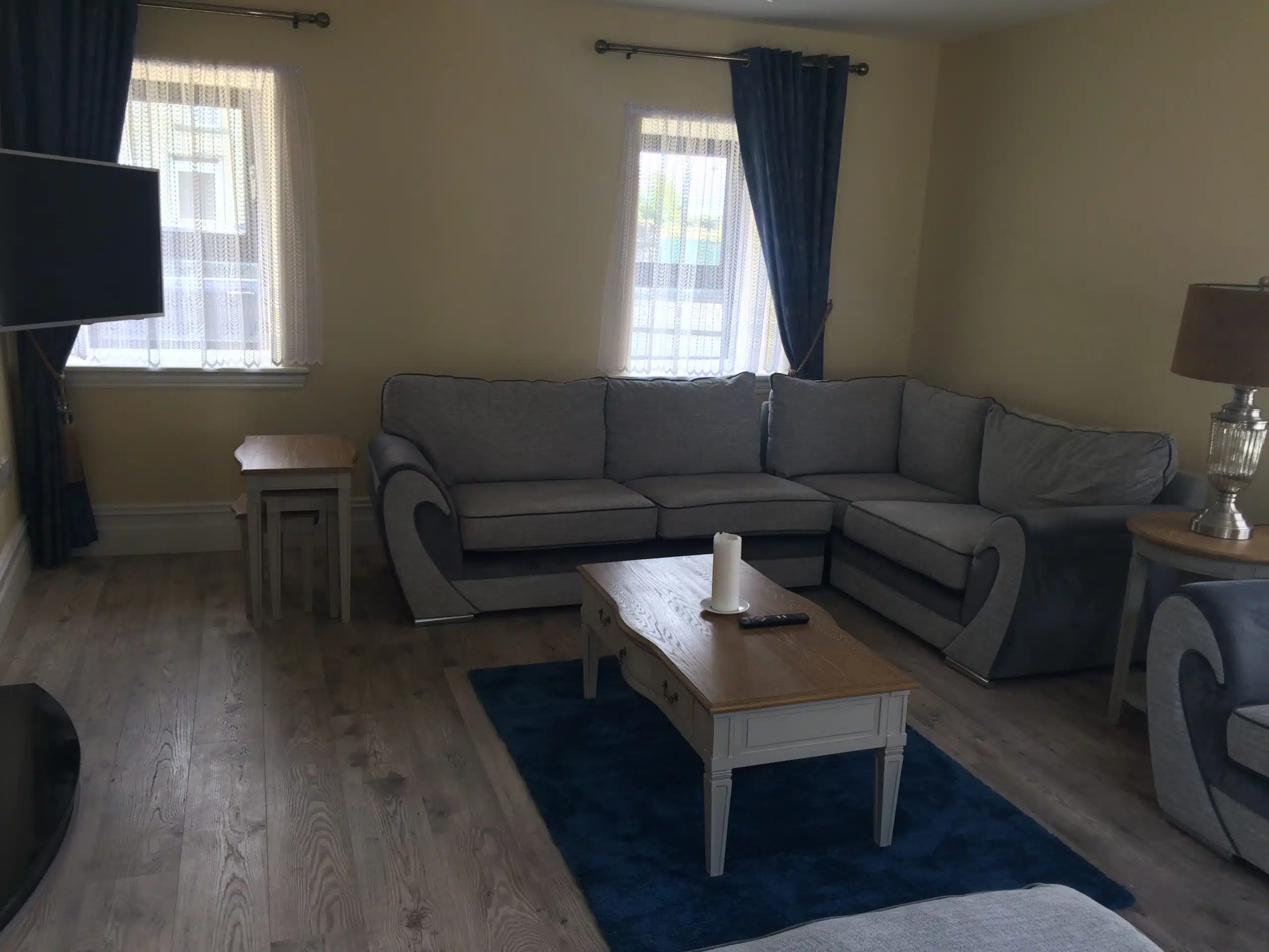 A living room with a grey sectional sofa, a wooden coffee table holding a candle and remote, a blue rug, two windows with sheer curtains, dark blue drapes, and a floor lamp on a side table. Wood flooring throughout.