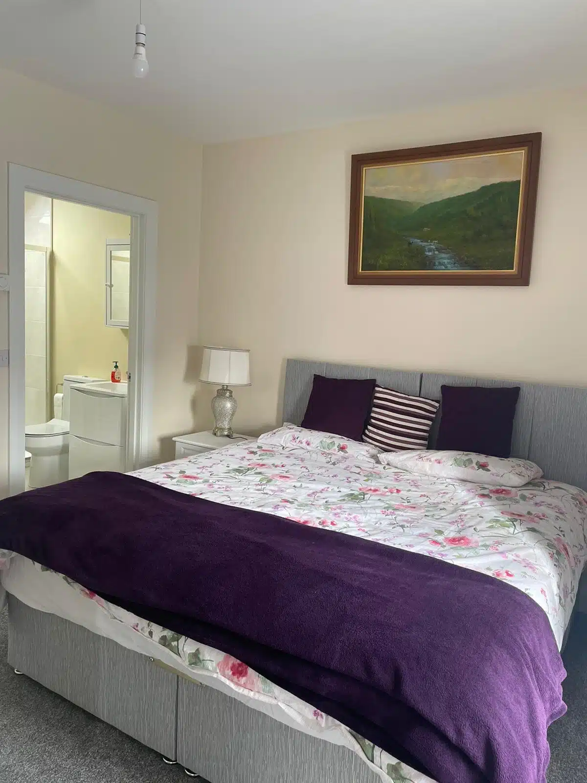 A neatly made bed with floral bedding and a purple blanket sits in a cosy bedroom. There are two purple pillows, a striped cushion, a bedside lamp, and a framed landscape painting above the bed. A bathroom is visible through an open door.