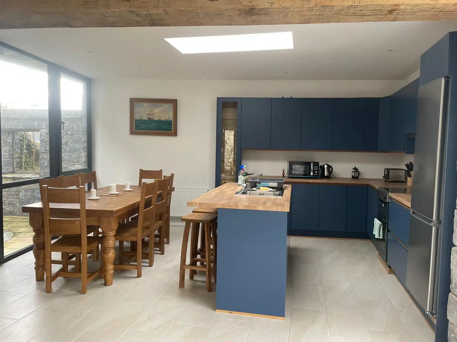 Modern kitchen and dining area with blue cupboards, wooden worktops, stainless steel appliances, and a wooden dining table with six chairs. A large window and skylight provide natural light. A painting hangs on the white wall.