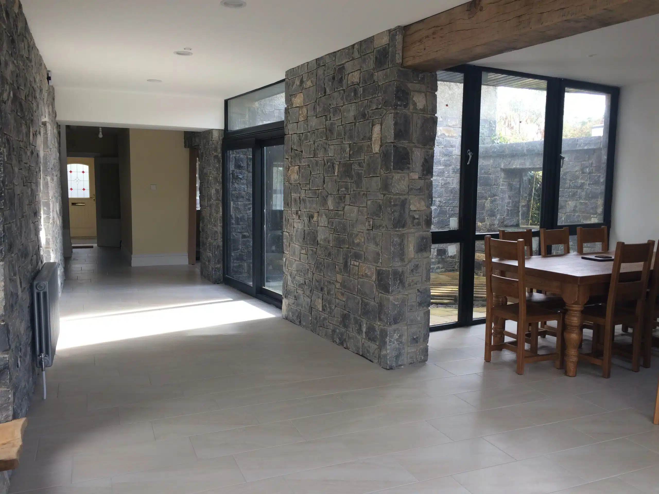 A modern dining area with stone walls, large windows, a wooden dining table with chairs, and tiled floors. Sunlight streams in through the windows, creating a bright and airy space.
