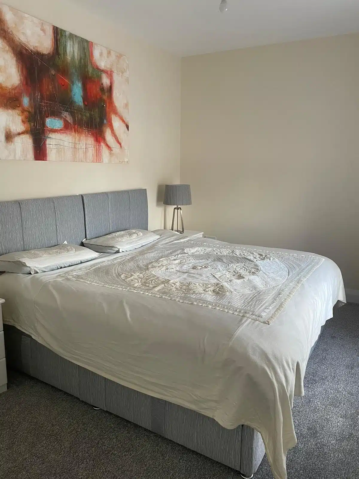 A neatly made bed with a light-coloured patterned bedspread sits in a modern, minimal bedroom. A grey headboard, two pillows, a bedside table with a lamp, and an abstract artwork decorate the room.