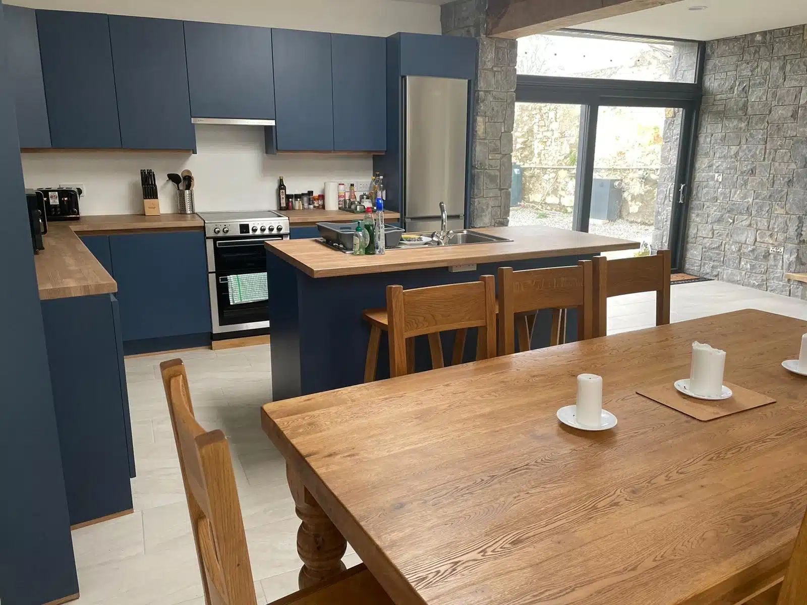 Modern kitchen with blue cupboards, wooden worktops, and a central island with a sink. A wooden dining table with four chairs and three white candles is in the foreground. Large windows and stone walls provide natural light.