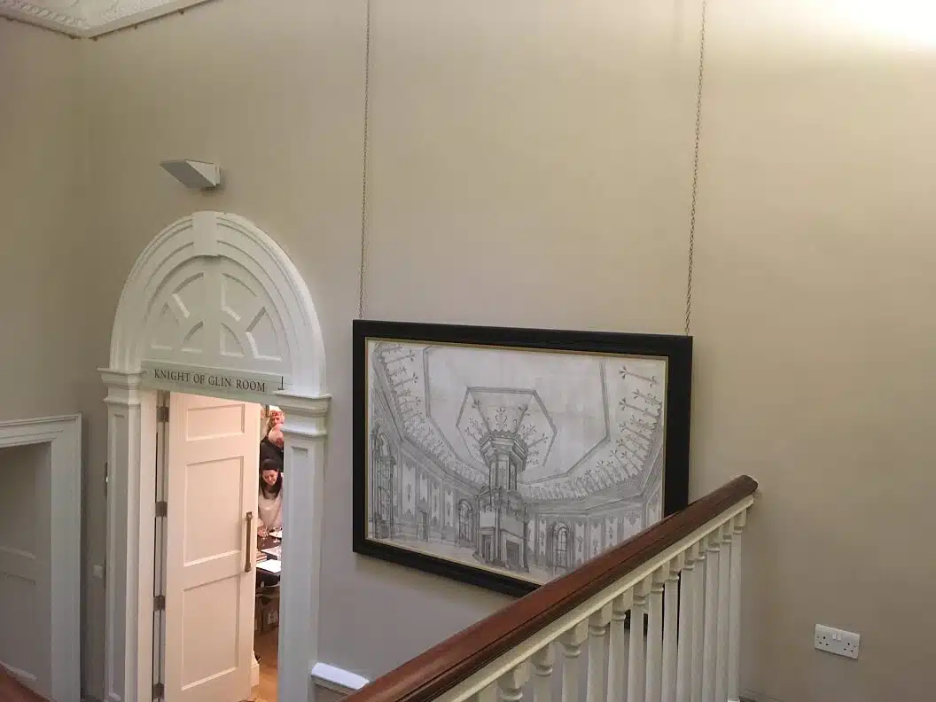 A staircase with a white bannister leads to a corridor. A large framed architectural drawing hangs on the wall. To the left, an open door labelled "Knight of Glin Room" reveals two people inside, one waving.