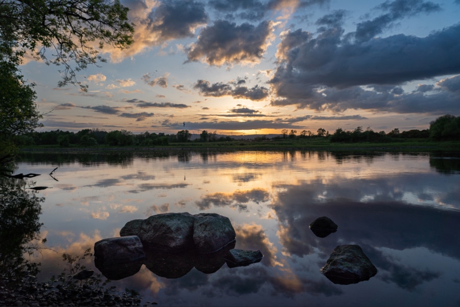 A tranquil lake at sunset with rocks in the foreground, trees along the shore, and colourful clouds reflected in the calm water.