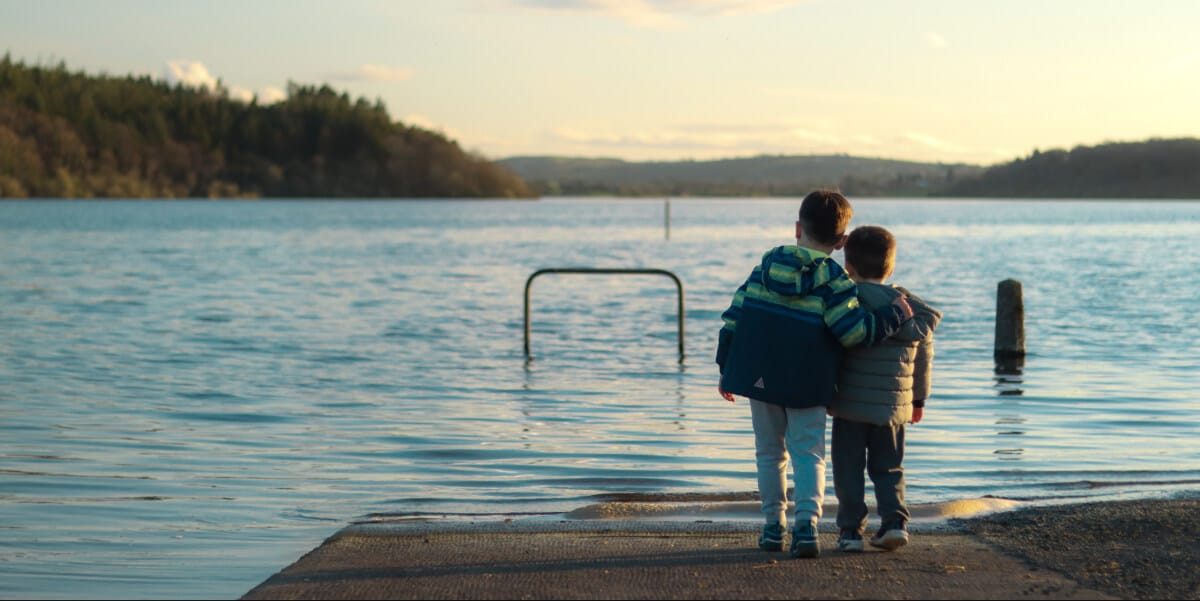 Two young children stand on a jetty by a calm lake, with one child’s arm around the other’s shoulder. Trees and hills are visible in the background under a clear, soft evening sky.