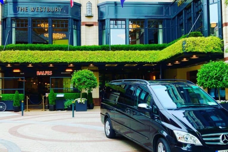 A black van is parked in front of The Westbury hotel entrance, with green hedges, potted plants, and large windows above the doorway. The Balfe’s restaurant sign is visible near the entrance.