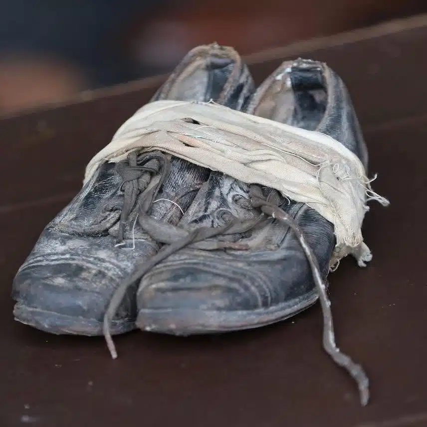 A pair of old, worn-out black shoes with frayed laces are wrapped together with a tattered white cloth, sitting on a dark surface.