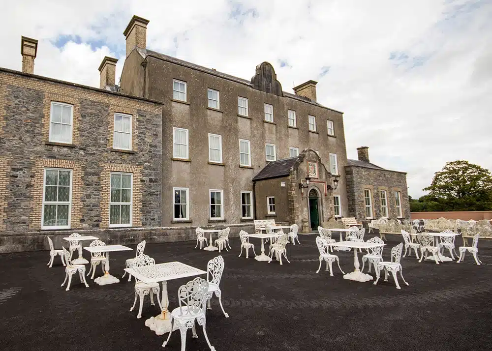 An old, grey stone building with large windows has an outdoor terrace filled with white metal tables and chairs, under a cloudy sky.