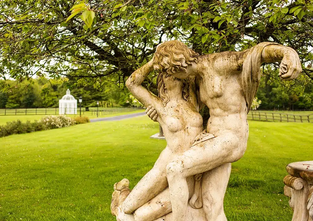 A weathered stone statue of two intertwined figures sits on a lush green lawn beneath a leafy tree, with a white gazebo and wooden fence visible in the background.