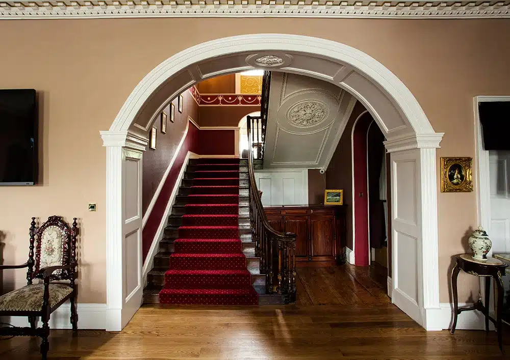 A grand staircase with a red carpet runner is framed by a large white archway in an elegant hallway with ornate mouldings, wooden floors, antique furniture, and artwork on the walls.