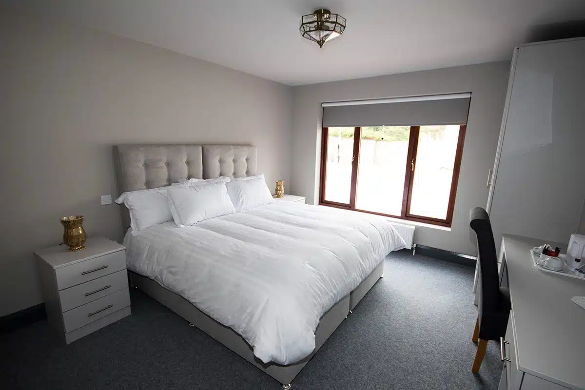 A modern, tidy bedroom with a large bed covered in white linen, two bedside tables with gold vases, a window with a blind, a white desk, and a black chair on grey carpeted flooring.