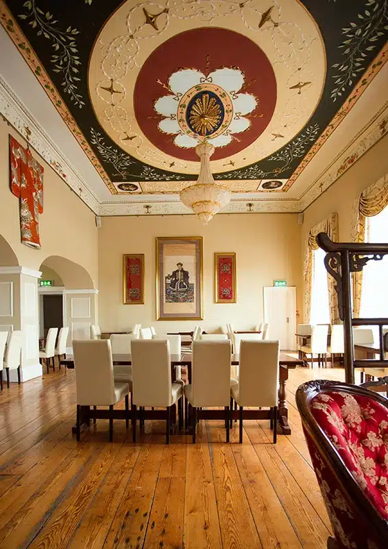 An elegant dining room with a long table and white chairs, ornate chandelier, decorative ceiling, wooden floor, and Asian-inspired artwork and decor on the walls. Natural light enters through large windows with draped curtains.