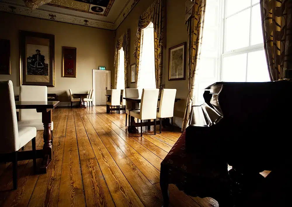 A large room with polished wooden floors, white cushioned chairs around tables, tall windows with patterned curtains, framed artwork on the walls, and a grand piano in the foreground. Sunlight streams through the windows.