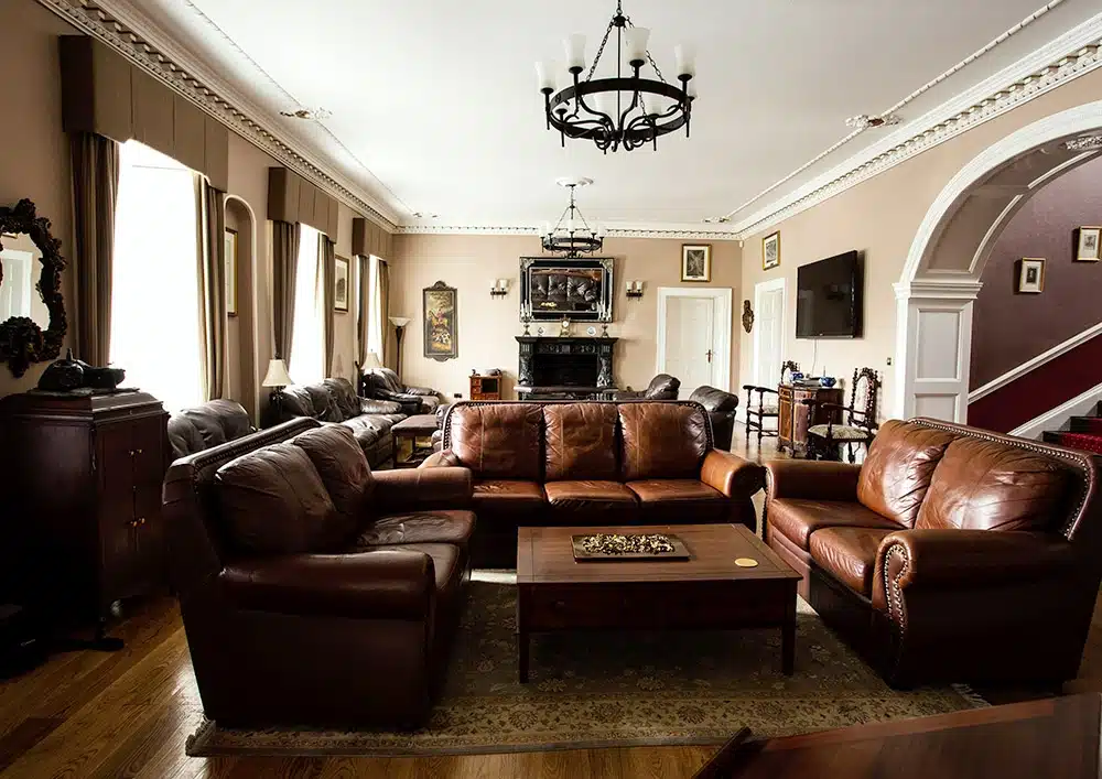 A spacious lounge with brown leather sofas around a wooden coffee table, wooden floors, a fireplace, wall-mounted TV, and large windows with curtains letting in natural light.