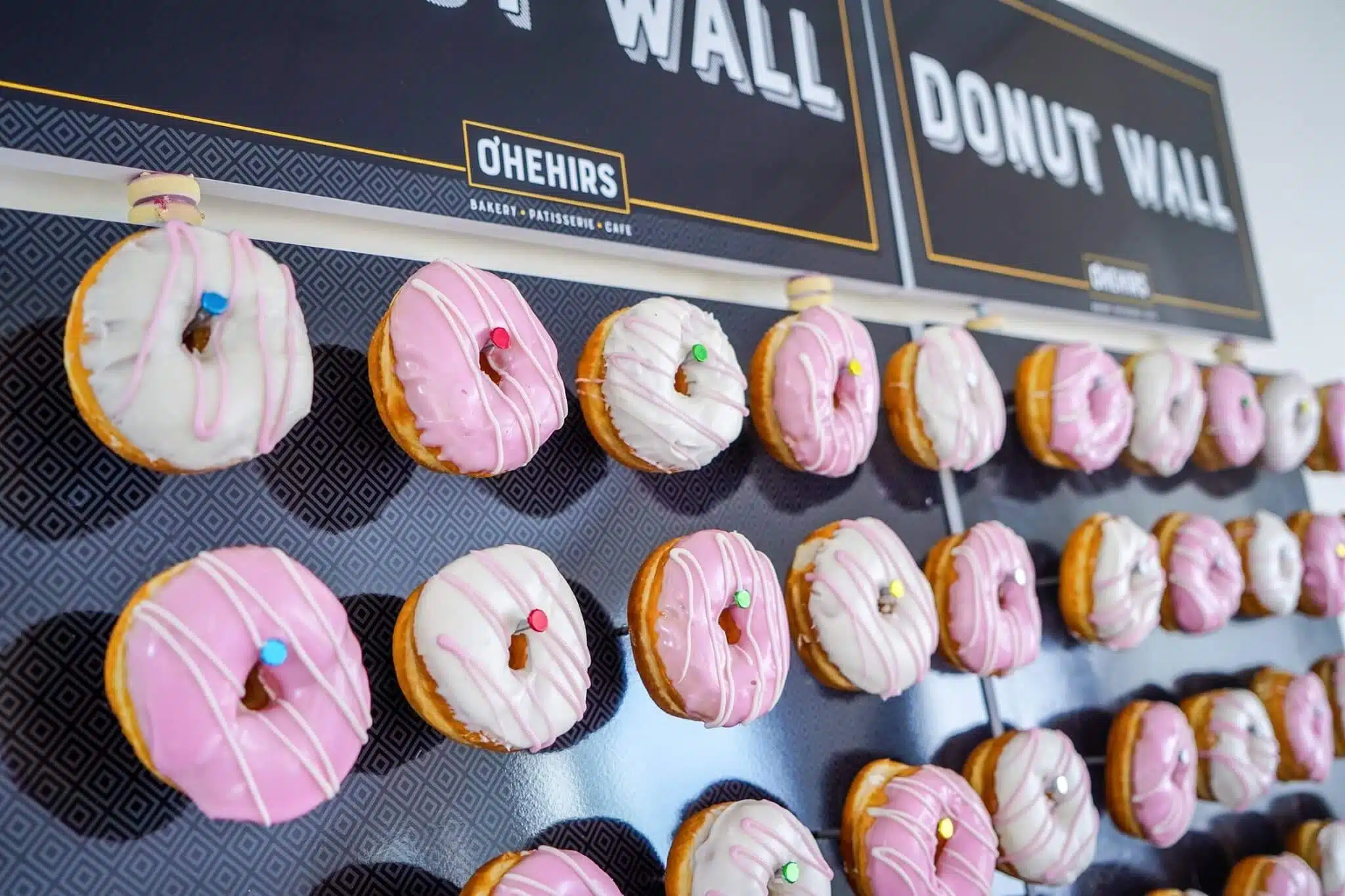 A display wall labelled "Doughnut Wall" holds rows of pink and white frosted doughnuts, each topped with a colourful chocolate sweet, attached by pegs for people to grab.