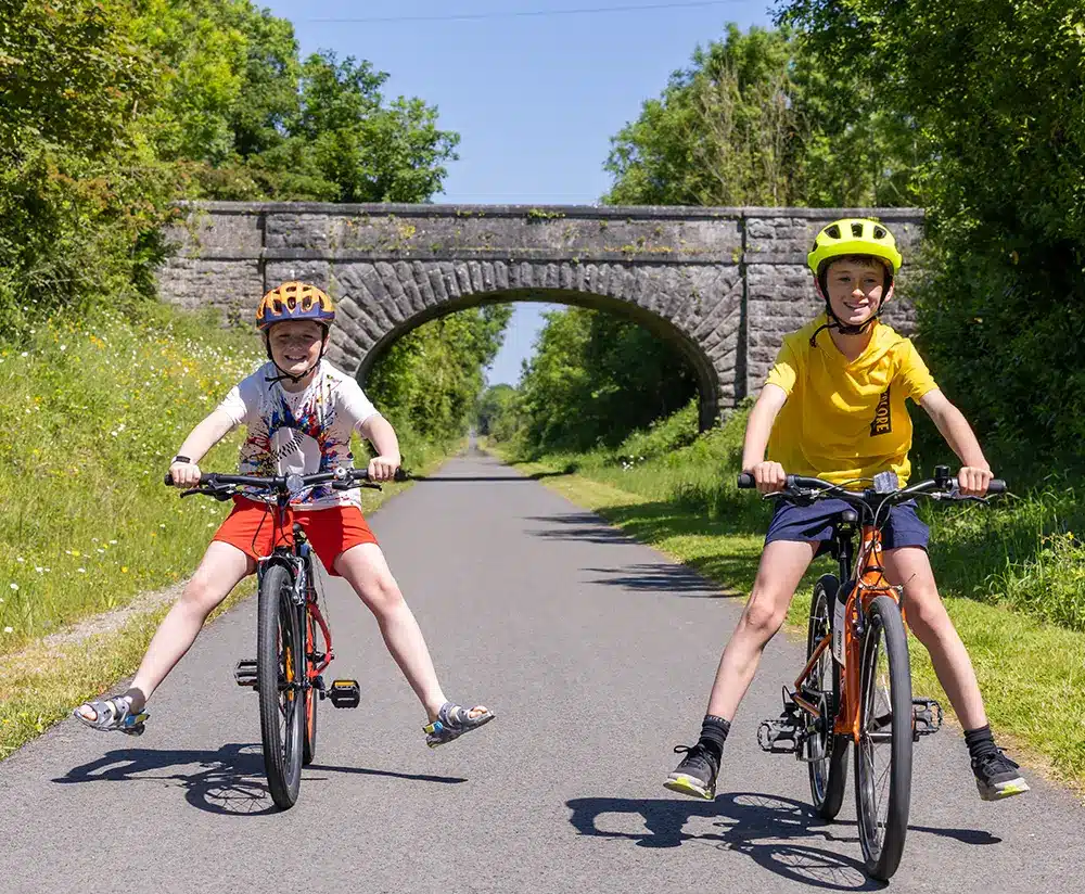 Two children wearing helmets ride bikes on a sunny, paved path with green trees and grass. One child wears red shorts and stretches their legs out; a stone bridge arches over the road in the background.