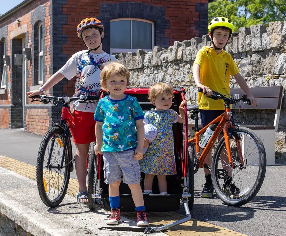 Four children are at a railway platform on a sunny day. Two older boys stand beside bikes, while two younger children, a boy and a girl, sit in a bike trailer. There’s a stone wall and brick building in the background.