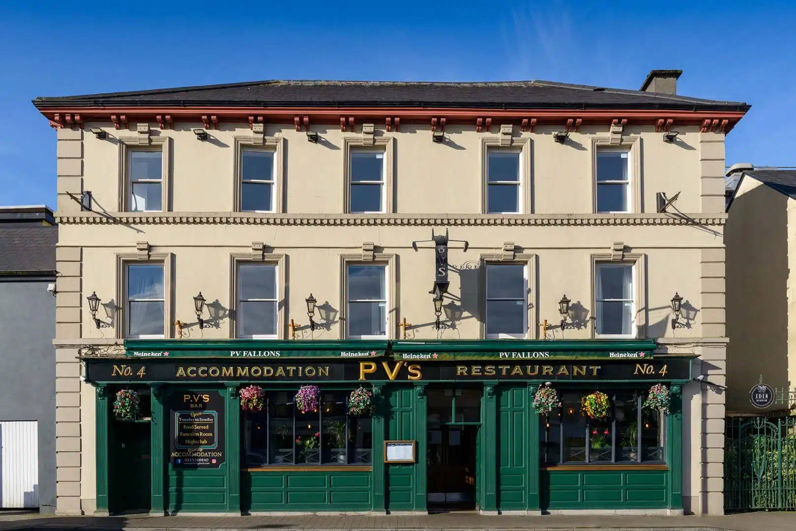A cream-coloured, three-storey building with green doors and window frames houses "P. Fallon's PV'S Restaurant & Accommodation." Hanging flower baskets decorate the facade under clear blue skies.