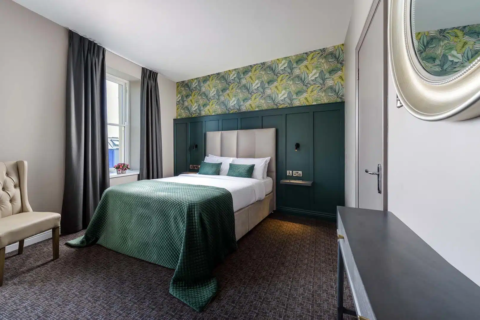 A modern hotel room with a double bed, green bedding, a padded headboard, patterned feature wall, a window with dark curtains, an upholstered chair, a wall mirror, and a dark desk.