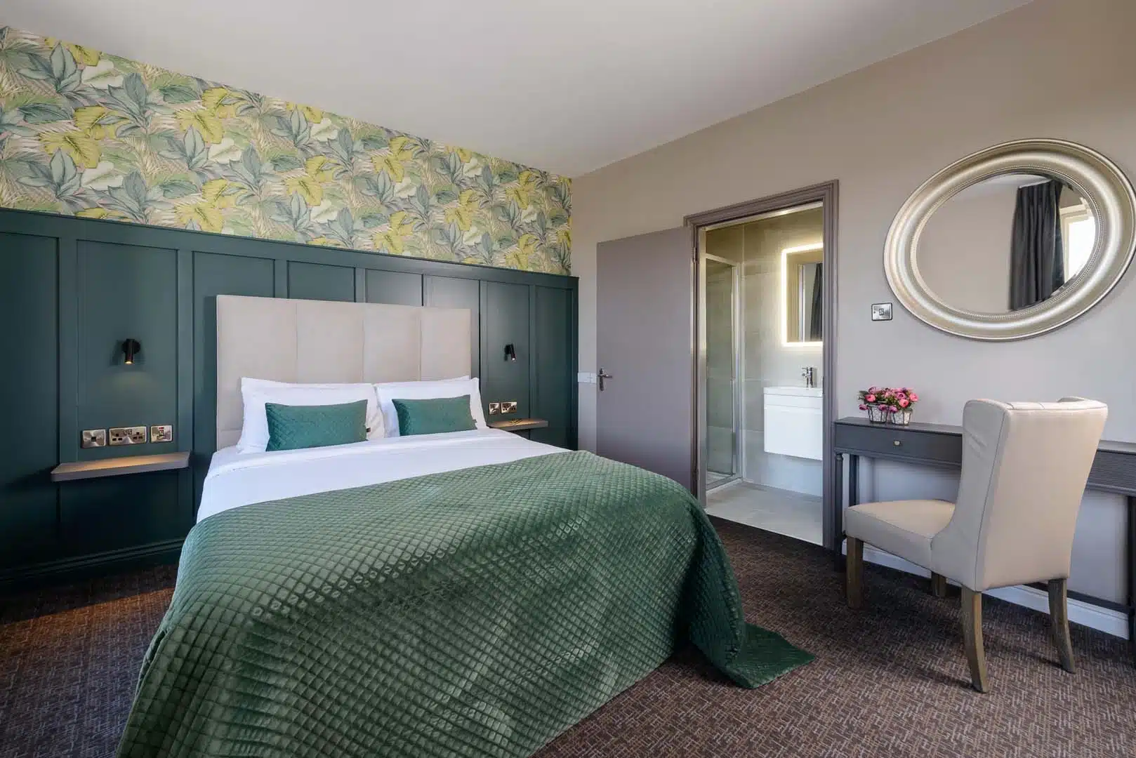 A modern hotel room with a double bed featuring green bedding and pillows, a green accent wall with floral wallpaper, a desk with a round mirror, a beige chair, and an open door to a bathroom.