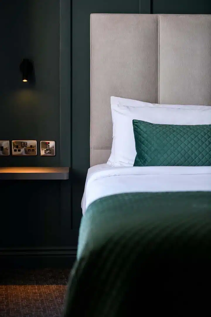 A modern hotel room features a neatly made bed with a green quilted blanket and matching pillow, white sheets, a beige headboard, a floating bedside table with sockets, and a wall-mounted reading light.