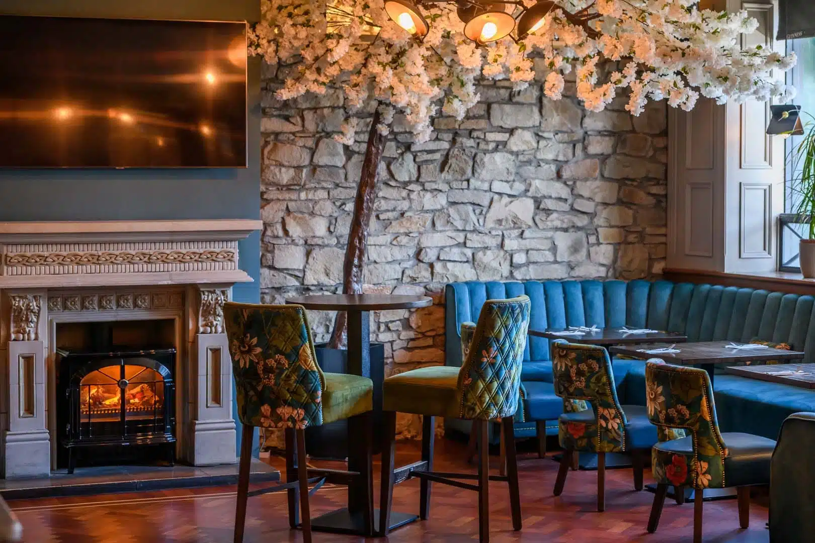 A cosy restaurant with stone walls, a fireplace, blue velvet seating, floral upholstered chairs, and a tree with white blossoms next to a window, creating a warm and inviting atmosphere.