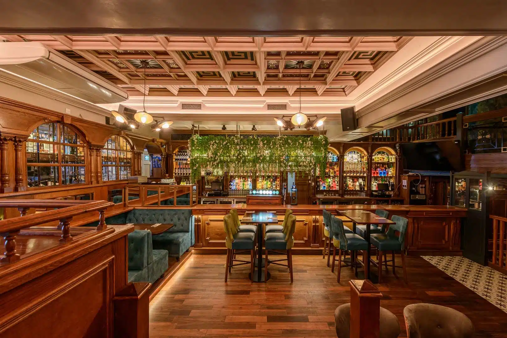 Interior of a stylish bar with wooden décor, stained glass accents, blue upholstered seating, a central bar area, hanging greenery, and warm ambient lighting. The atmosphere is inviting and elegant.