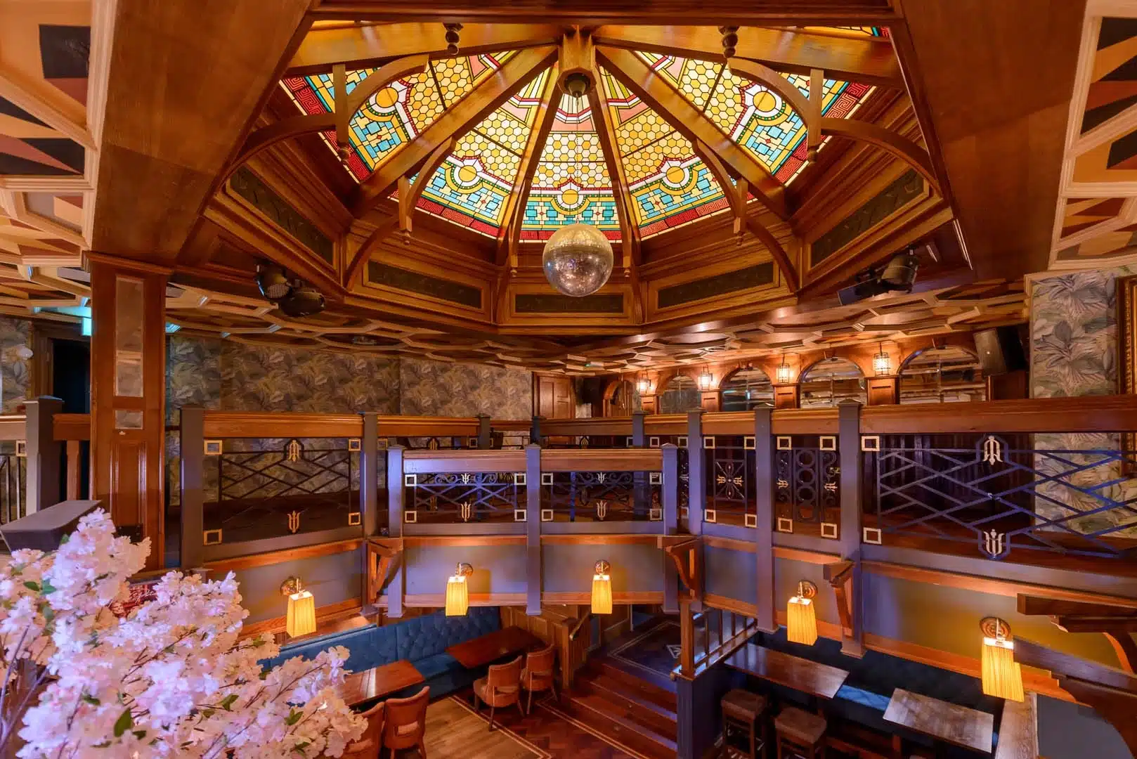 A stylish restaurant interior featuring wooden decor, warm lighting, a mezzanine level with booths, and a colourful stained glass dome ceiling with a disco ball at the centre. Tables and chairs are arranged below.