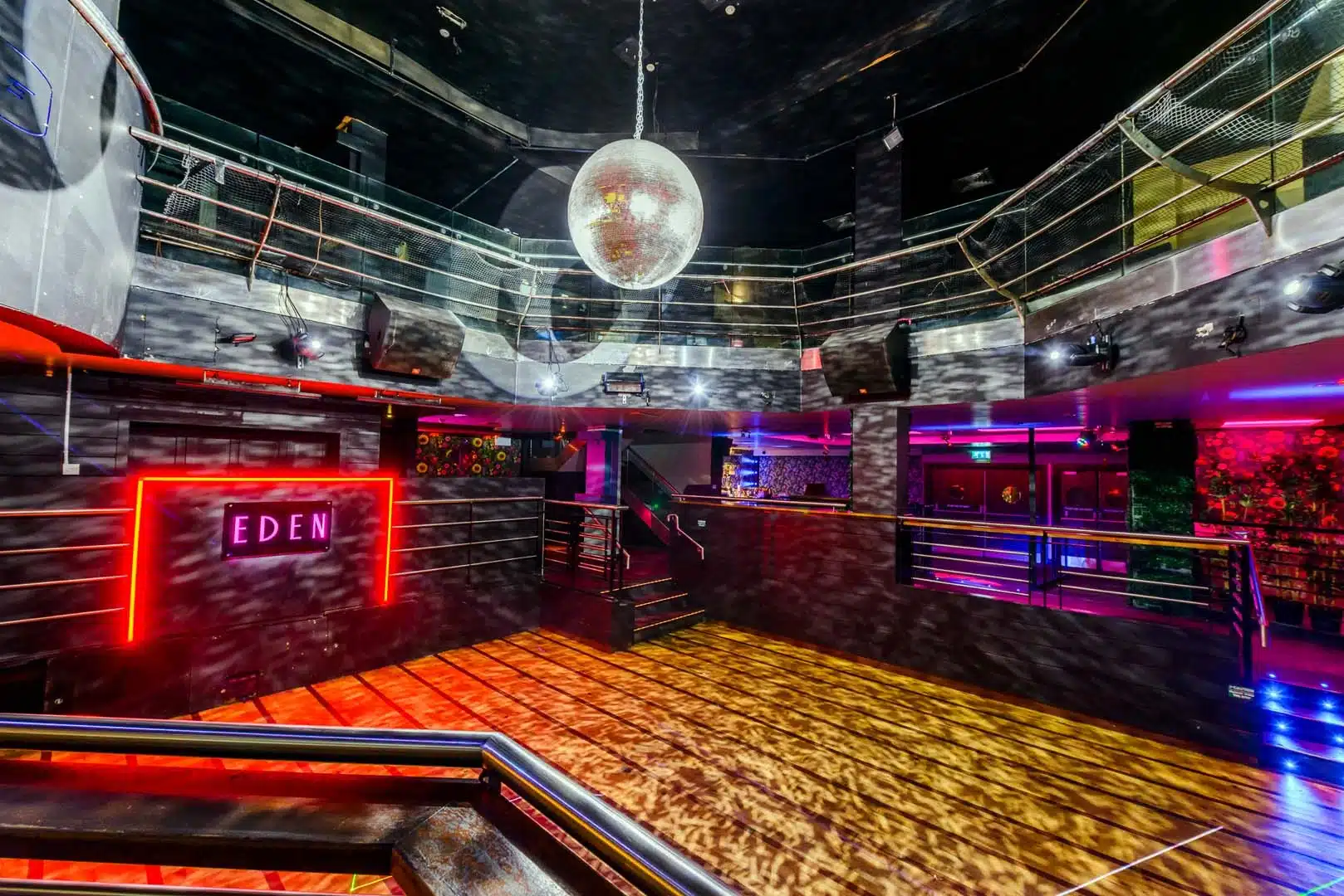 A modern nightclub interior with a large disco ball hanging from the ceiling, colourful neon and spot lights, a dance floor, and a glowing red sign that reads "EDEN" on the wall.