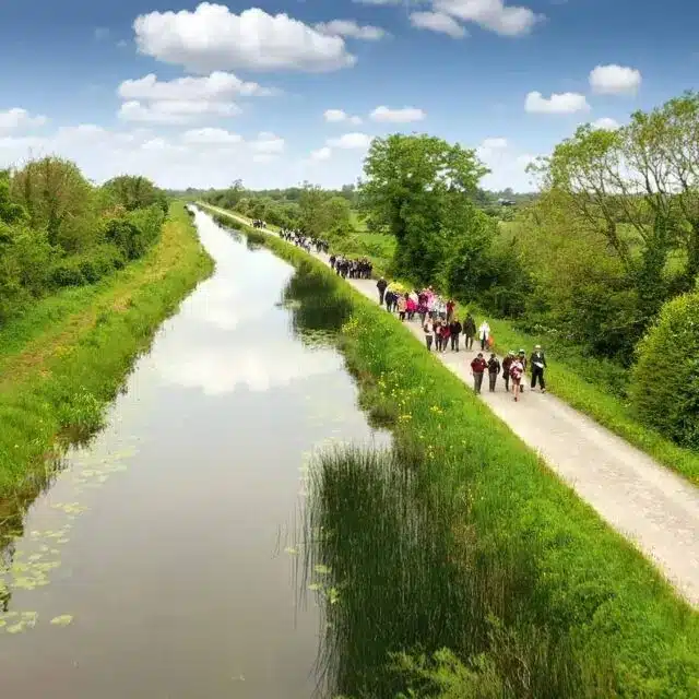 A group of people walk along a gravel path beside a calm canal, surrounded by green grass, trees, and bushes under a blue sky with scattered clouds.
