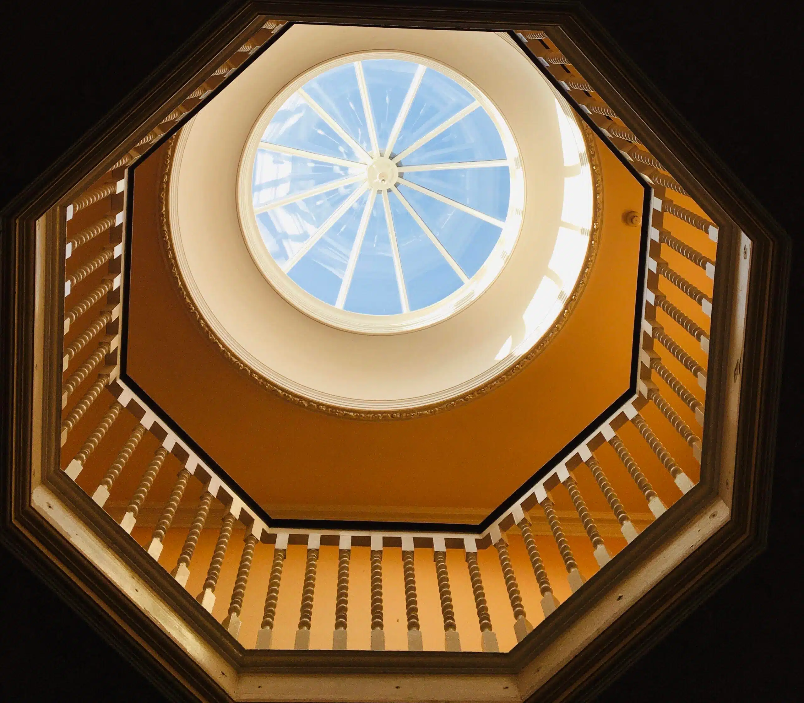 View looking up through an octagonal staircase with wooden railings, revealing a large, round skylight at the top letting in bright natural light.